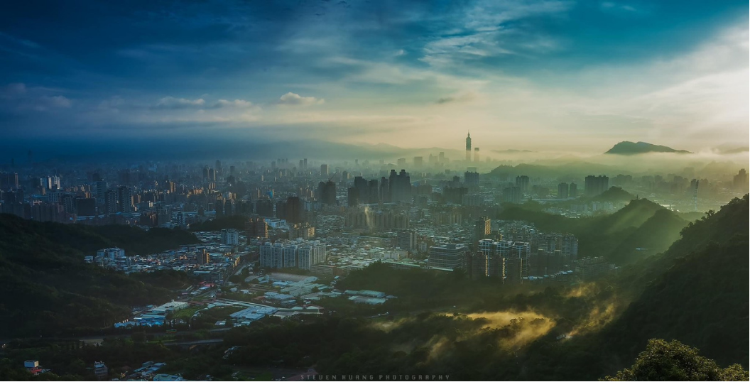 從山地高處俯瞰臺北盆地與城市景觀，臺北101位於遠方，周圍可見山稜、城市與天空。